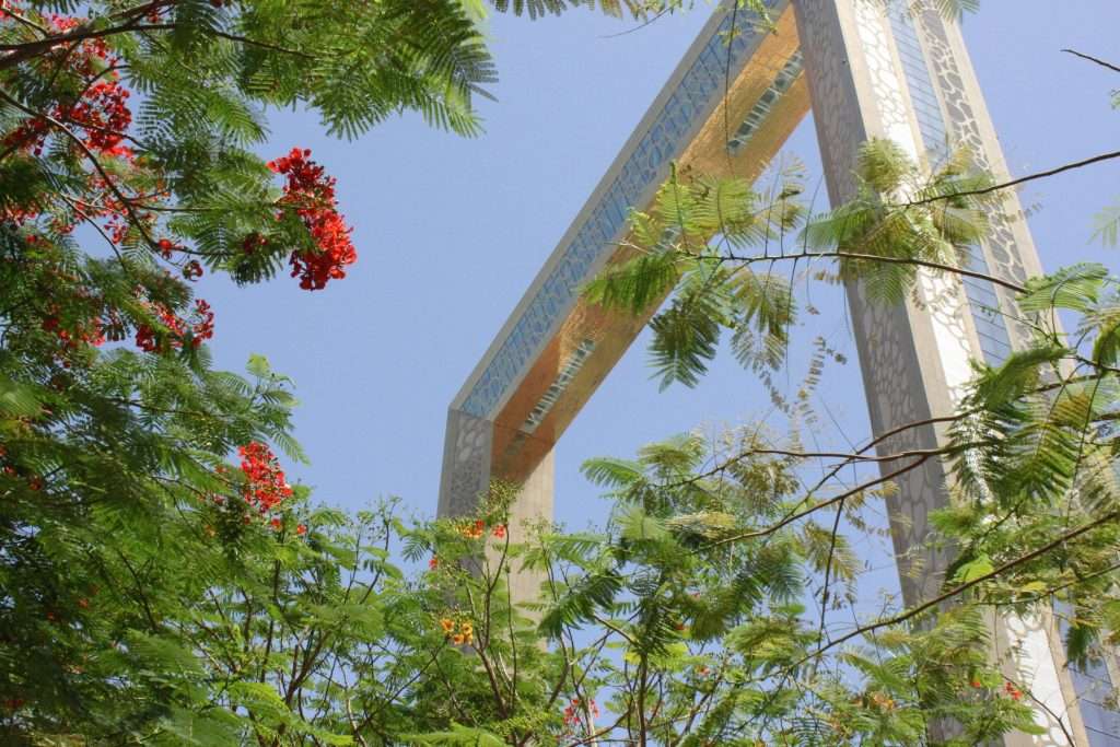 Looking up at a large rectangular architectural frame with intricate designs, surrounded by green leafy branches and clusters of red flowers against a clear blue sky—a view that could easily be featured in The Ultimate Autism-Friendly Guide to Dubai.